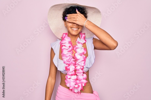 Young beautiful african american tourist woman wearing bikini and hawaiian lei flowers smiling and laughing with hand on face covering eyes for surprise. Blind concept.