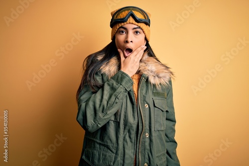 Young beautiful hispanic woman wearing ski glasses and coat for winter weather Looking fascinated with disbelief, surprise and amazed expression with hands on chin