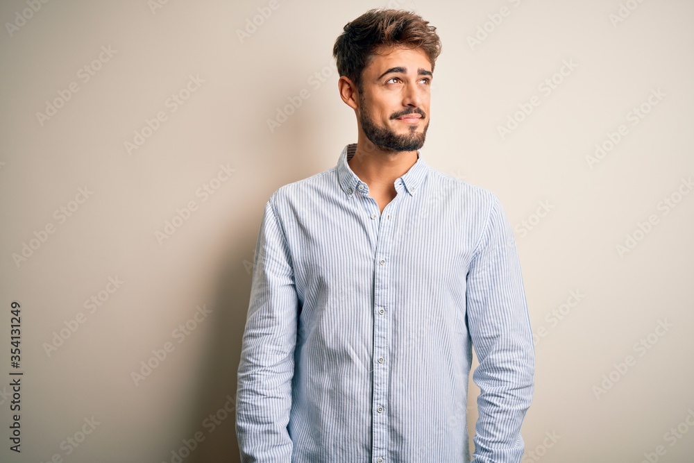 Young handsome man with beard wearing striped shirt standing over white background smiling looking to the side and staring away thinking. Young handsome man with beard wearing striped shirt standing over white background smiling looking to the side and staring away thinking.