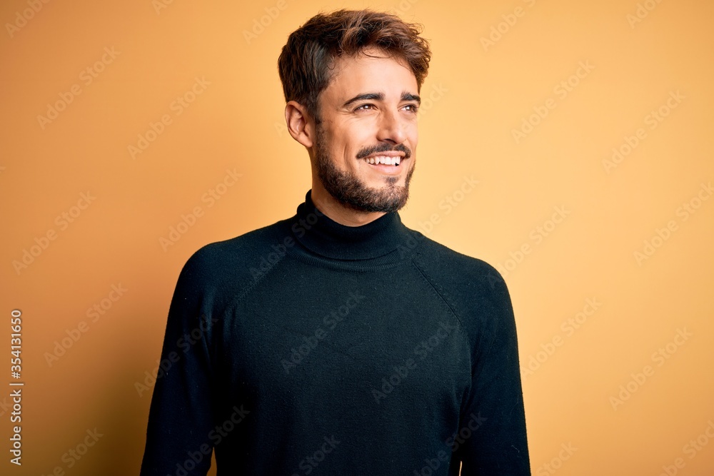 Young handsome man with beard wearing turtleneck sweater standing over yellow background looking away to side with smile on face, natural expression. Laughing confident. Young handsome man with beard wearing turtleneck sweater standing over yellow background looking away to side with smile on face, natural expression. Laughing confident.