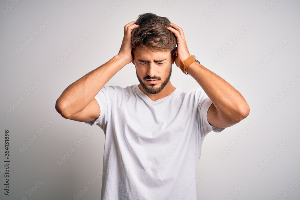 Young handsome man with beard wearing casual t-shirt standing over white background suffering from headache desperate and stressed because pain and migraine. Hands on head. Young handsome man with beard wearing casual t-shirt standing over white background suffering from headache desperate and stressed because pain and migraine. Hands on head.