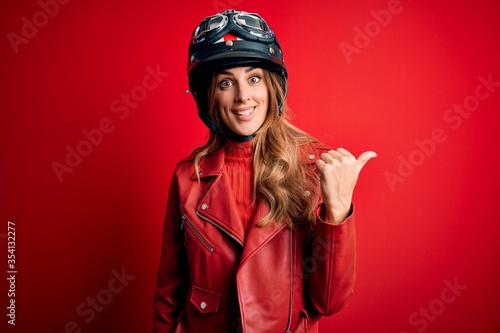 Young beautiful brunette motrocyclist woman wearing moto helmet over red background smiling with happy face looking and pointing to the side with thumb up. © Krakenimages.com