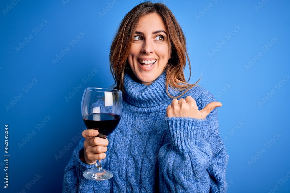 Young beautiful brunette woman drinking glass of red wine over isolated blue background pointing and showing with thumb up to the side with happy face smiling Young beautiful brunette woman drinking glass of red wine over isolated blue background pointing and showing with thumb up to the side with happy face smiling
