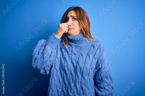 Young beautiful brunette woman wearing casual turtleneck sweater over blue background smelling something stinky and disgusting, intolerable smell, holding breath with fingers on nose. Bad smell