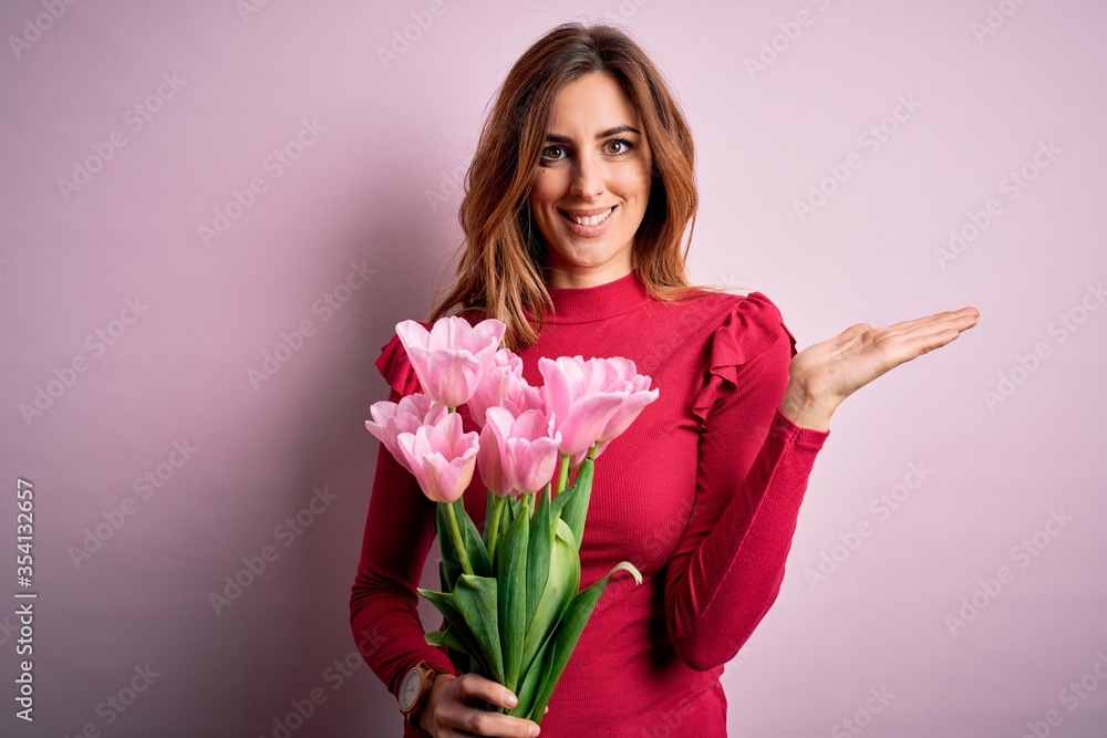 Young beautiful brunette woman holding bouquet of pink tulips over isolated background smiling cheerful presenting and pointing with palm of hand looking at the camera. Young beautiful brunette woman holding bouquet of pink tulips over isolated background smiling cheerful presenting and pointing with palm of hand looking at the camera.