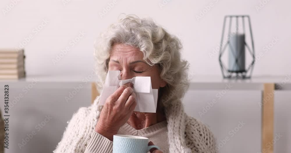 Head shot unhealthy senior old woman using paper tissues, wiping runny nose, drinking warming anti grippe beverage, sitting on sofa covered in knitted plaid, cold season illnesses, healthcare concept.