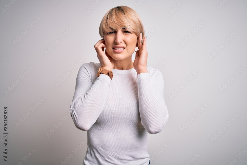 Young beautiful blonde woman with modern short hair hairstyle standing over isolated background covering ears with fingers with annoyed expression for the noise of loud music. Deaf concept. Young beautiful blonde woman with modern short hair hairstyle standing over isolated background covering ears with fingers with annoyed expression for the noise of loud music. Deaf concept.