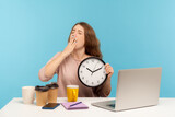 Tired office employee working late hours. Exhausted woman surrounded by coffee cups yawning and holding clock, feeling fatigue, stress of overtime job. indoor studio shot isolated on blue background