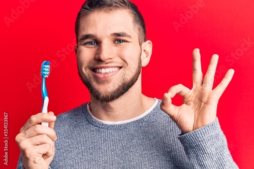 Young handsome man holding toothbrush doing ok sign with fingers, smiling friendly gesturing excellent symbol