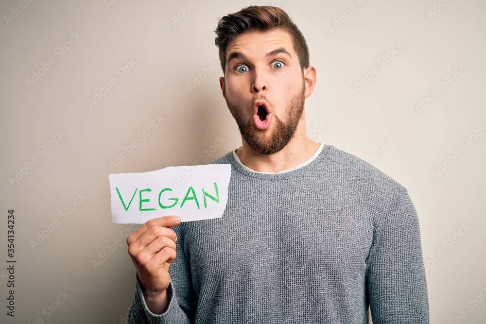 Young blond veggie man with beard and blue eyes holding paper with vegan message scared in shock with a surprise face, afraid and excited with fear expression Young blond veggie man with beard and blue eyes holding paper with vegan message scared in shock with a surprise face, afraid and excited with fear expression