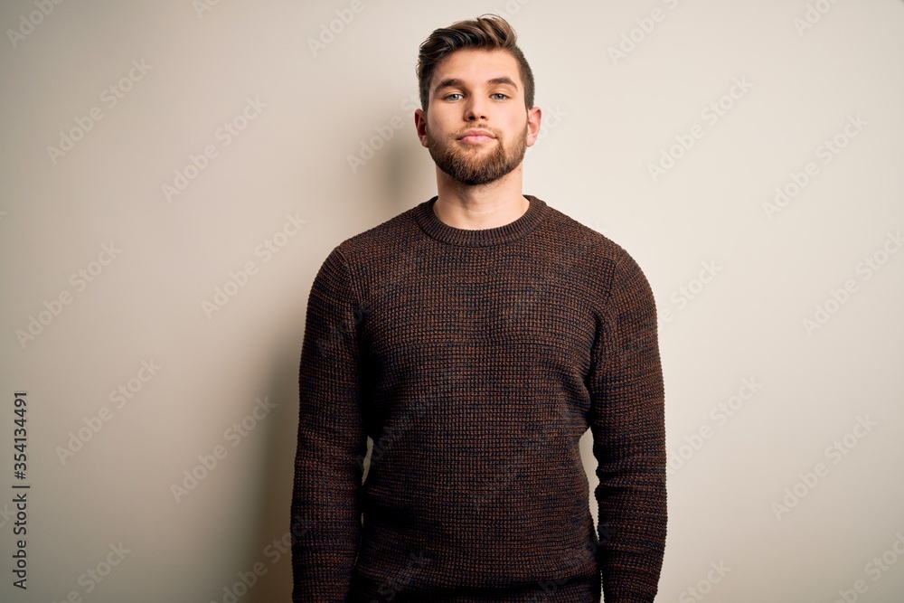 Young handsome blond man with beard and blue eyes wearing casual sweater standing with serious expression on face. Simple and natural looking at the camera. Young handsome blond man with beard and blue eyes wearing casual sweater standing with serious expression on face. Simple and natural looking at the camera.