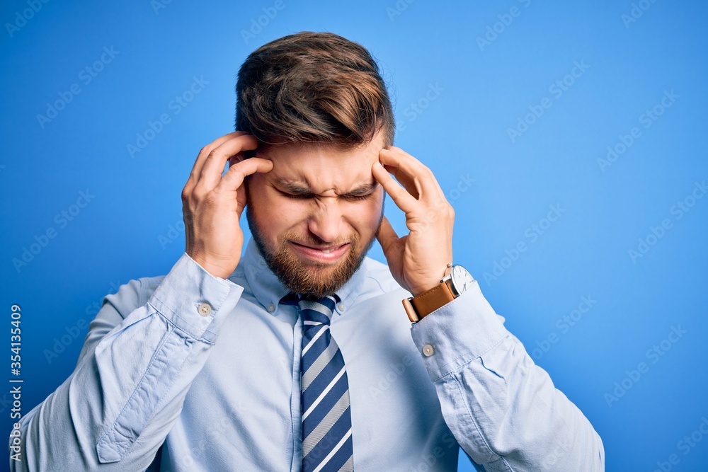 Young blond businessman with beard and blue eyes wearing elegant shirt and tie standing with hand on head for pain in head because stress. Suffering migraine. Young blond businessman with beard and blue eyes wearing elegant shirt and tie standing with hand on head for pain in head because stress. Suffering migraine.