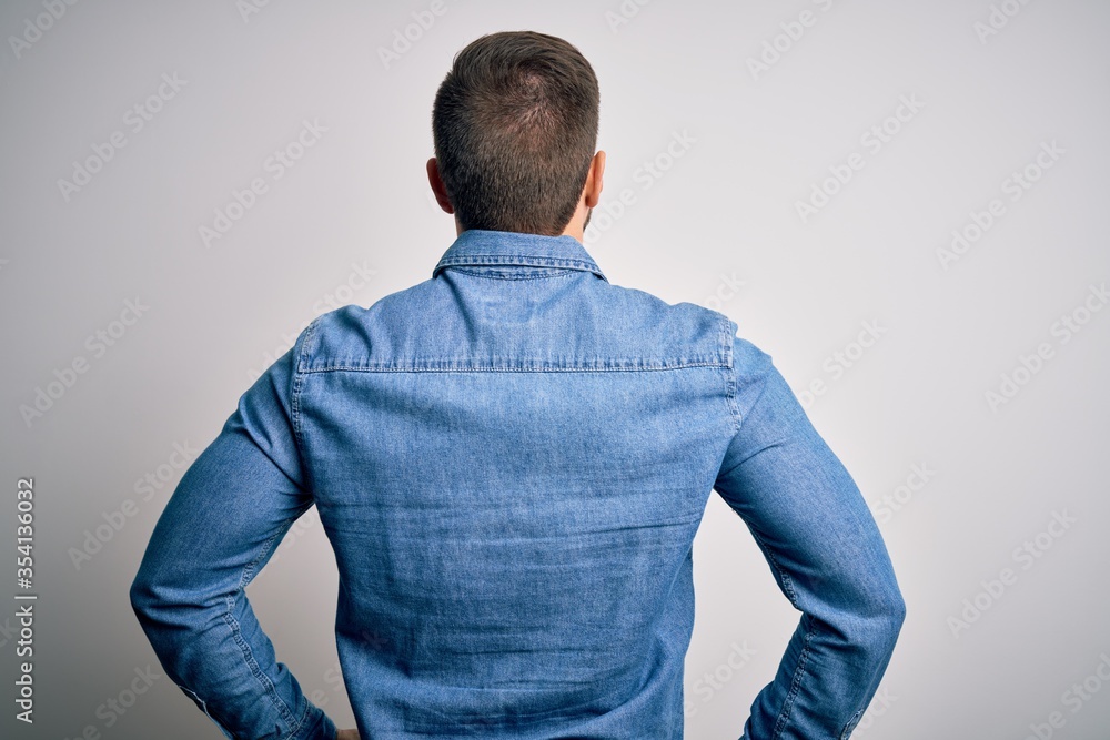 Young handsome blond man with beard and blue eyes wearing casual denim shirt standing backwards looking away with arms on body Young handsome blond man with beard and blue eyes wearing casual denim shirt standing backwards looking away with arms on body