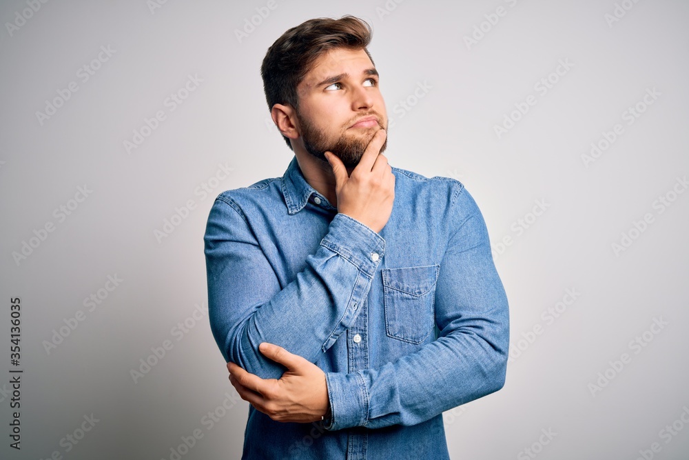 Young handsome blond man with beard and blue eyes wearing casual denim shirt with hand on chin thinking about question, pensive expression. Smiling with thoughtful face. Doubt concept. Young handsome blond man with beard and blue eyes wearing casual denim shirt with hand on chin thinking about question, pensive expression. Smiling with thoughtful face. Doubt concept.