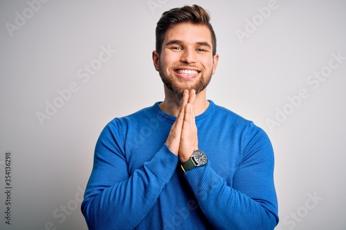 Young handsome blond man with beard and blue eyes wearing casual sweater praying with hands together asking for forgiveness smiling confident.