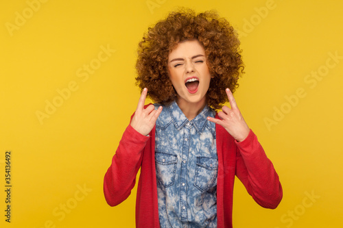 Let's rock! Portrait of crazy woman with curly hair in casual outfit showing rock and roll sign and roaring angrily, looking aggressive disobedient. indoor studio shot isolated on yellow background