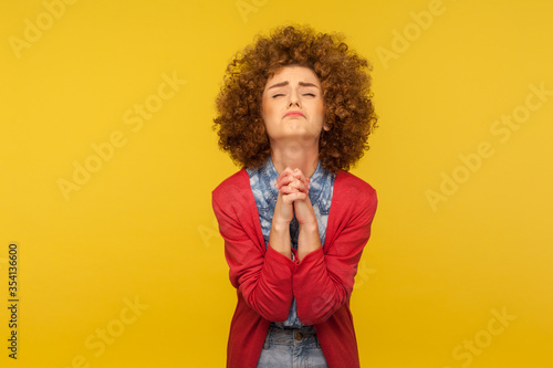 Please, I'm begging! Portrait of upset worried woman with curly hair looking up with imploring desperate grimace, praying to god asking for help. indoor studio shot isolated on yellow background photo