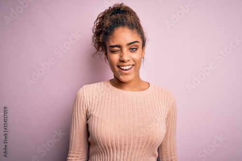 Young beautiful african american girl wearing casual sweater standing over pink background winking looking at the camera with sexy expression, cheerful and happy face.