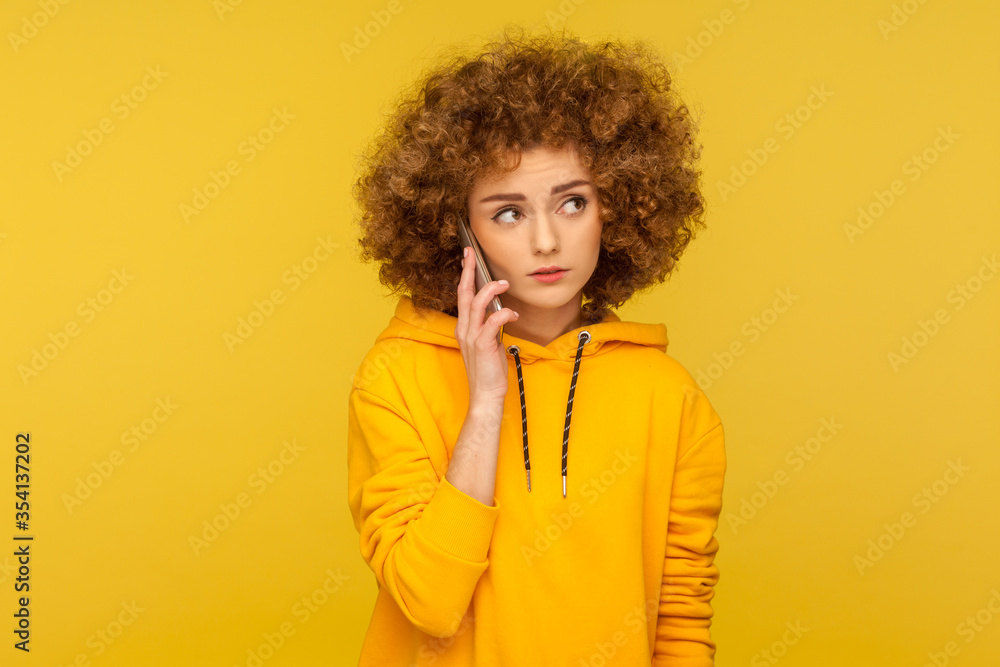 Portrait of upset curly-haired woman in urban style hoodie calling friend, talking on smartphone with serious unhappy expression, discussing bad news. indoor studio shot isolated on yellow background Portrait of upset curly-haired woman in urban style hoodie calling friend, talking on smartphone with serious unhappy expression, discussing bad news. indoor studio shot isolated on yellow background