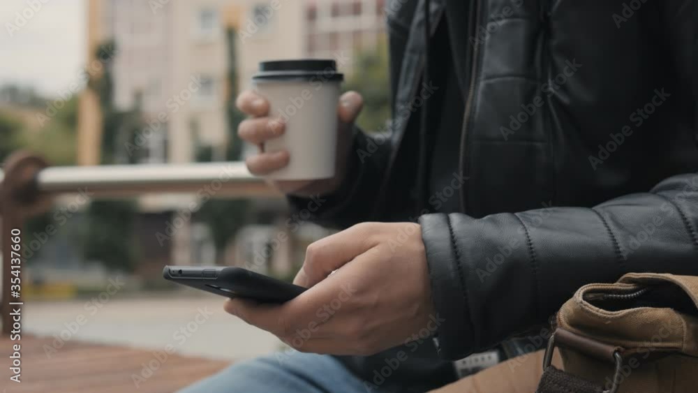 Man sitting on bench in the city centre drinking coffee and use a smartphone Man sitting on bench in the city centre drinking coffee and use a smartphone