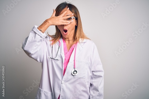 Middle age beautiful doctor woman wearing pink stethoscope over isolated white background peeking in shock covering face and eyes with hand, looking through fingers with embarrassed expression.