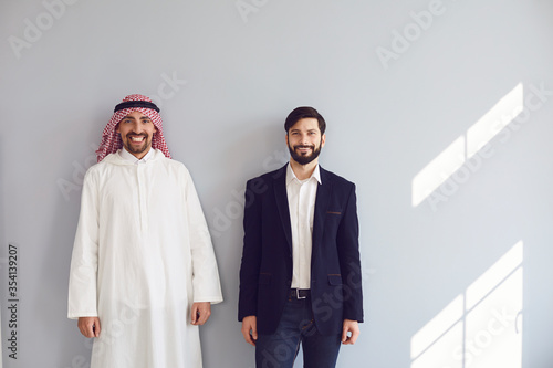 Arab and european businessmen stand smiling at the gray background.