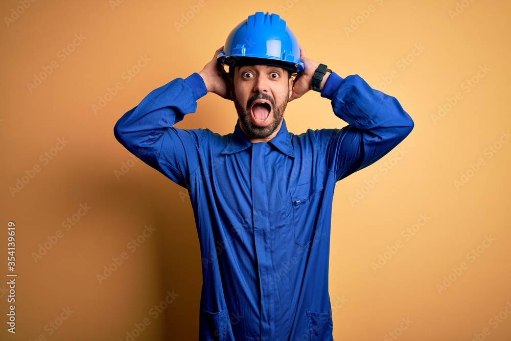Mechanic man with beard wearing blue uniform and safety helmet over yellow background Crazy and scared with hands on head, afraid and surprised of shock with open mouth