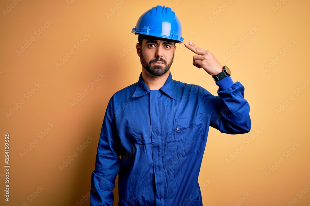 Mechanic man with beard wearing blue uniform and safety helmet over yellow background Shooting and killing oneself pointing hand and fingers to head like gun, suicide gesture.