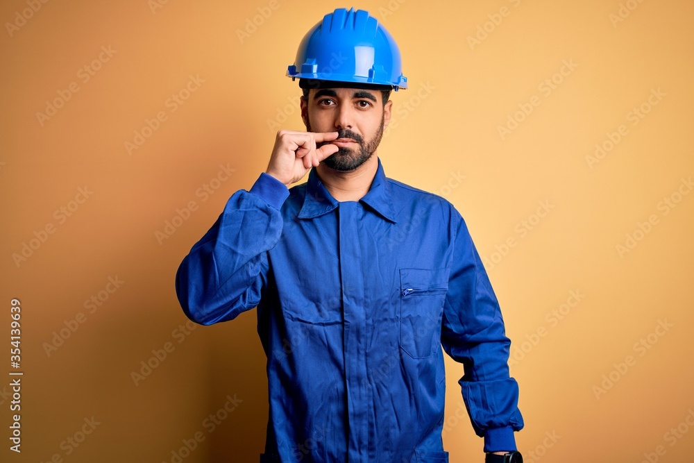 Mechanic man with beard wearing blue uniform and safety helmet over yellow background mouth and lips shut as zip with fingers. Secret and silent, taboo talking