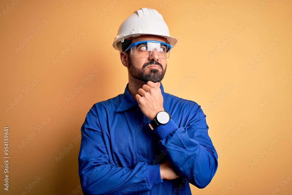Mechanic man with beard wearing blue uniform and safety glasses over yellow background with hand on chin thinking about question, pensive expression. Smiling with thoughtful face. Doubt concept.