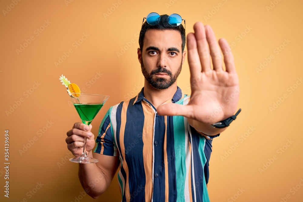 Young handsome tourist man with beard on vacation wearing summer shirt drinking cocktail with open hand doing stop sign with serious and confident expression, defense gesture Young handsome tourist man with beard on vacation wearing summer shirt drinking cocktail with open hand doing stop sign with serious and confident expression, defense gesture