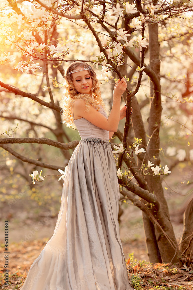 Beauty and fashion concept. Young beauty blonde with jewelry for hair, in a silver dress with a long train in the garden where magnolia trees bloom. Flares, bokeh effects. Beauty and fashion concept. Young beauty blonde with jewelry for hair, in a silver dress with a long train in the garden where magnolia trees bloom. Flares, bokeh effects.