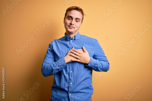 Young handsome redhead man wearing casual striped shirt over isolated yellow background smiling with hands on chest with closed eyes and grateful gesture on face. Health concept.