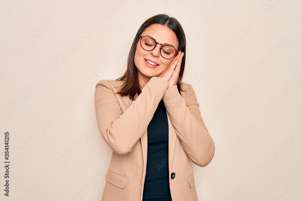 Young beautiful brunette businesswoman wearing jacket and glasses over white background sleeping tired dreaming and posing with hands together while smiling with closed eyes.