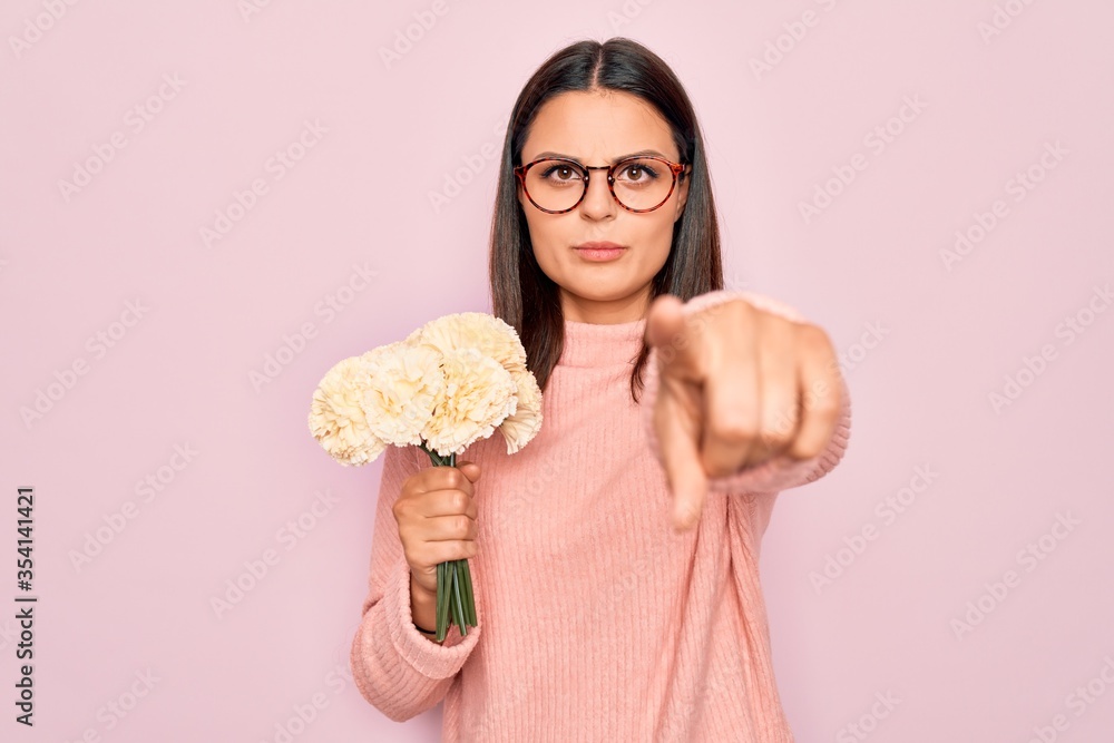 Beautiful brunette woman wearing glasses holding bouquet of flowers over pink background pointing with finger to the camera and to you, confident gesture looking serious