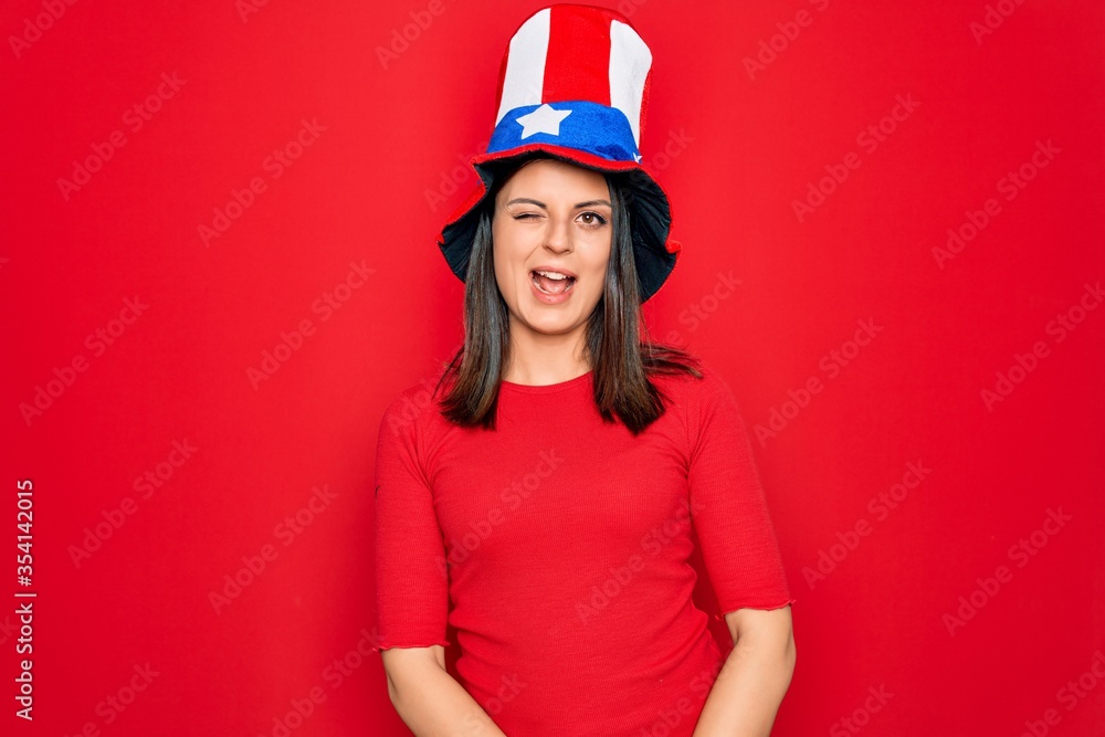 Young beautiful brunette woman wearing united states hat celebrating independence day winking looking at the camera with sexy expression, cheerful and happy face. Young beautiful brunette woman wearing united states hat celebrating independence day winking looking at the camera with sexy expression, cheerful and happy face.