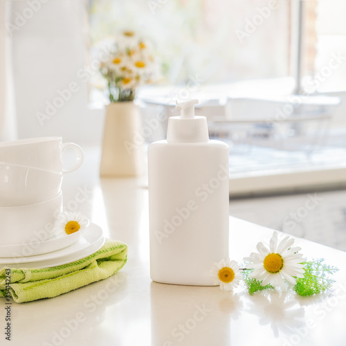 Eco friendly non-toxic cleaning dish soap with natural ingredients, chamomile flowers, clean white cups and plates on white kitchen table. Skincare for housekeeping. Bio organic cleaning supplies.