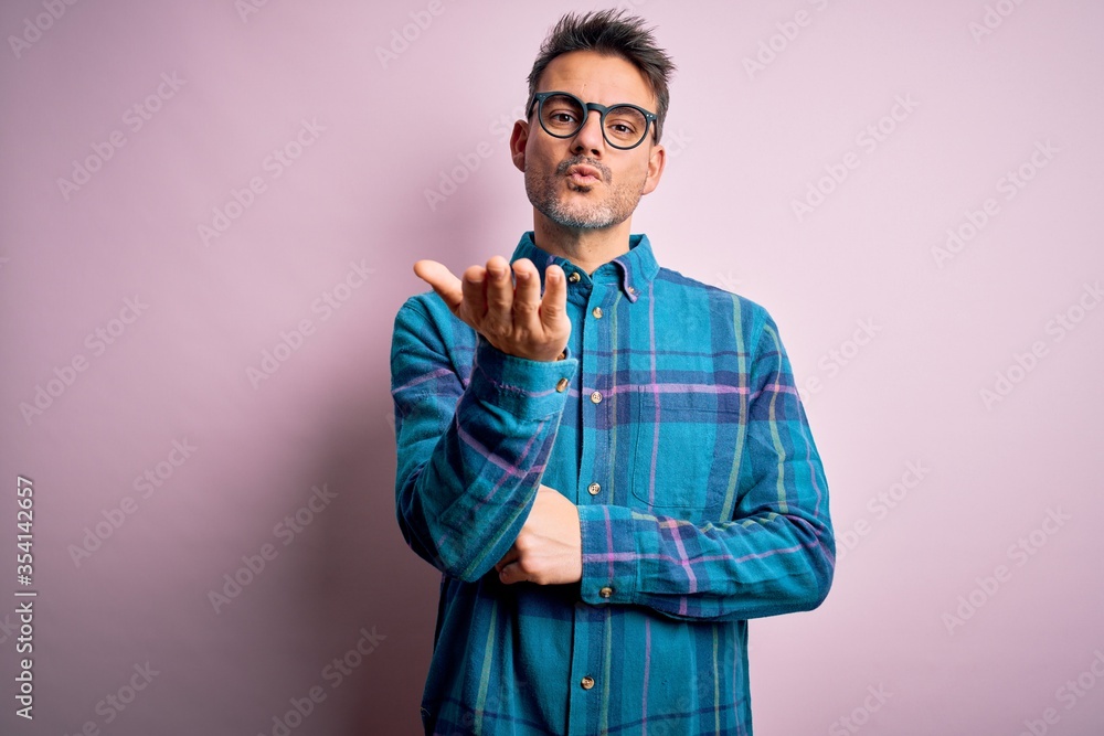 Young handsome man wearing casual shirt and glasses standing over isolated pink background looking at the camera blowing a kiss with hand on air being lovely and sexy. Love expression. Young handsome man wearing casual shirt and glasses standing over isolated pink background looking at the camera blowing a kiss with hand on air being lovely and sexy. Love expression.
