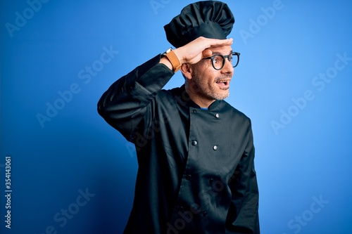 Young handsome chef man wearing cooker uniform and hat over isolated blue background very happy and smiling looking far away with hand over head. Searching concept.