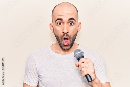 Young handsome bald man singing song using microphone scared and amazed with open mouth for surprise, disbelief face