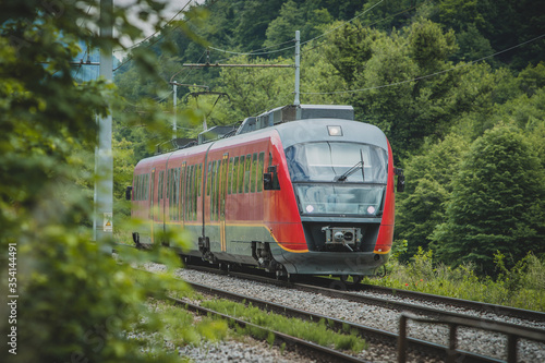 Red modern commuter train rushing on railway tracks through the green forest.