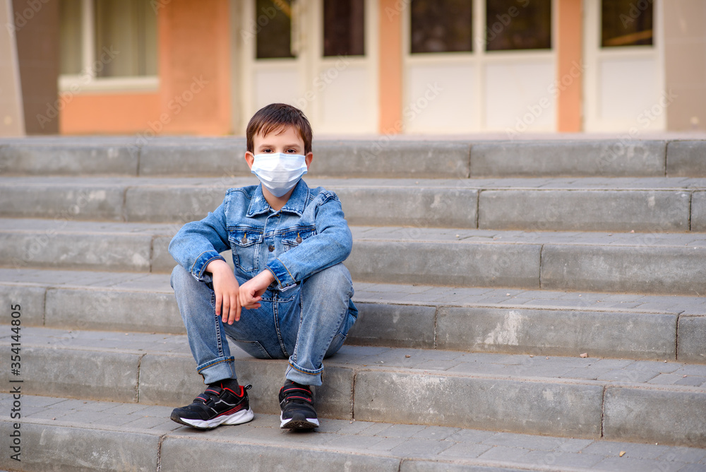 Schoolboy in protective mask sits on stairs outdoors in schoolyard. Pandemic coronavirus 2020.