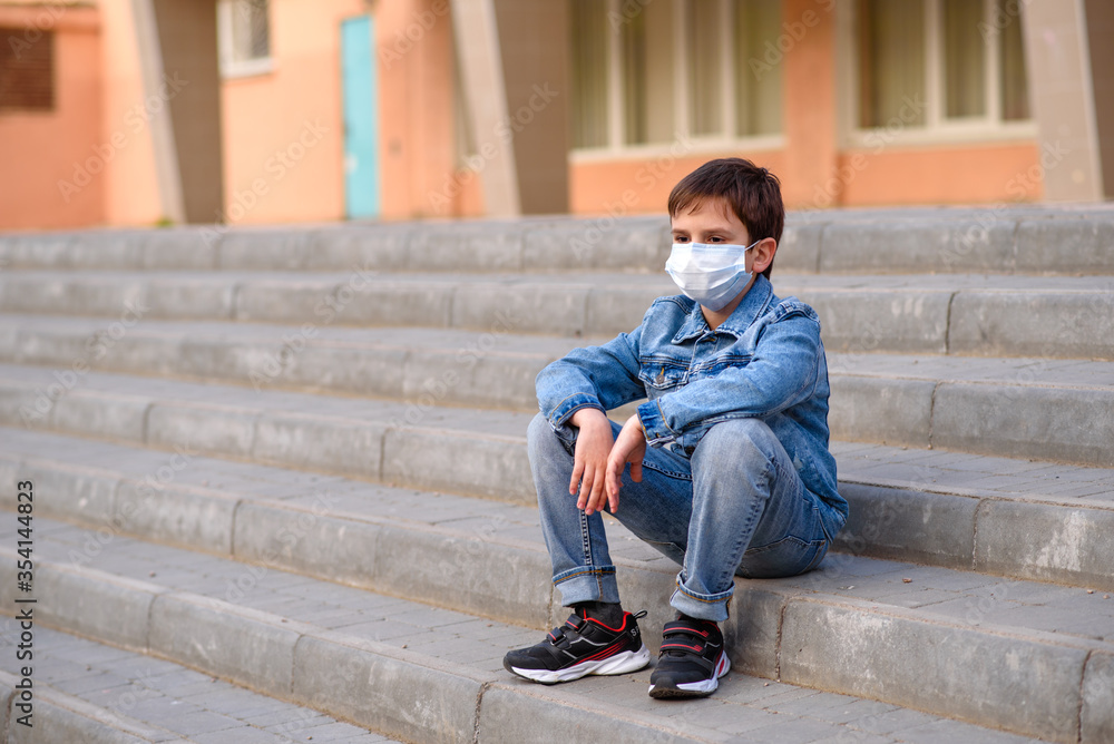 Schoolboy in protective mask in jeans clothes, black trainers, sits on stairs outdoors in schoolyard. Quarantine, Pandemic coronavirus 2020.
