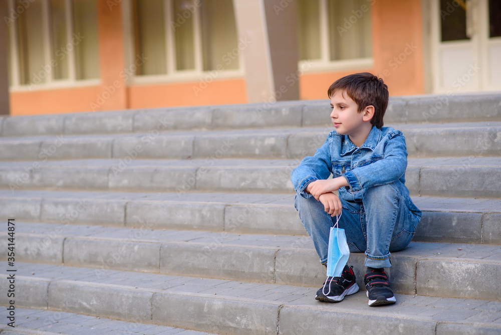 Smiling schoolboy sits on stairs outdoors in schoolyard with protective mask in hands. End of pandemic coronavirus 2020.End of quarantine covid-19 Smiling schoolboy sits on stairs outdoors in schoolyard with protective mask in hands. End of pandemic coronavirus 2020.End of quarantine covid-19