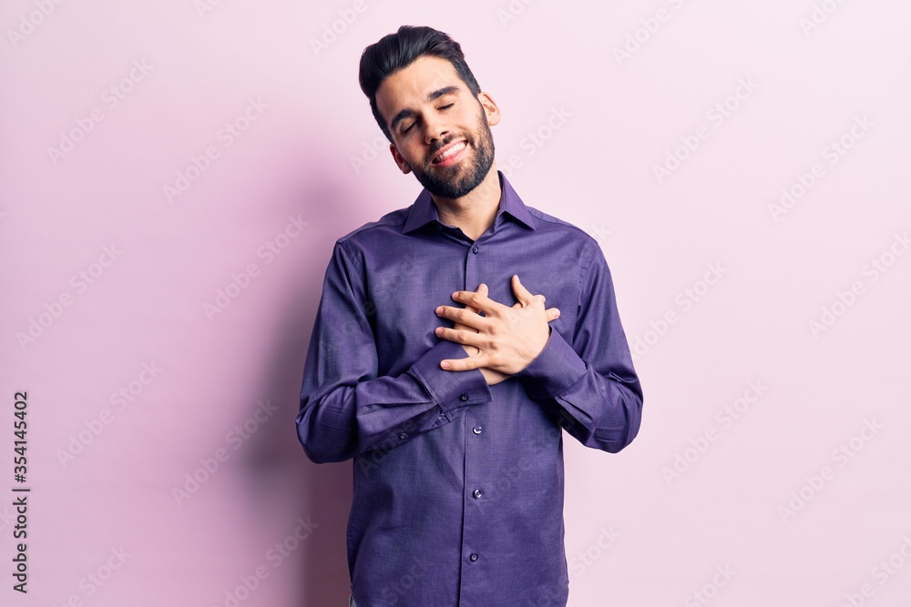 Young handsome man with beard wearing casual shirt smiling with hands on chest with closed eyes and grateful gesture on face. health concept. Young handsome man with beard wearing casual shirt smiling with hands on chest with closed eyes and grateful gesture on face. health concept.