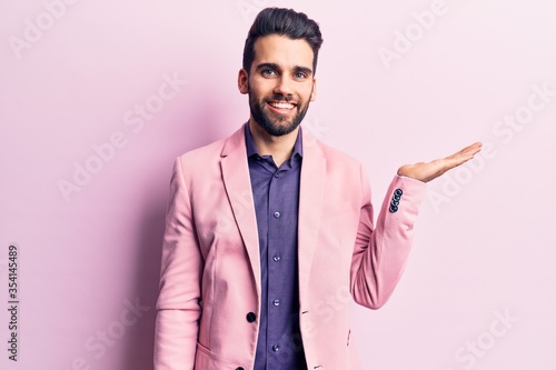 Young handsome man with beard wearing elegant jacket smiling cheerful presenting and pointing with palm of hand looking at the camera.