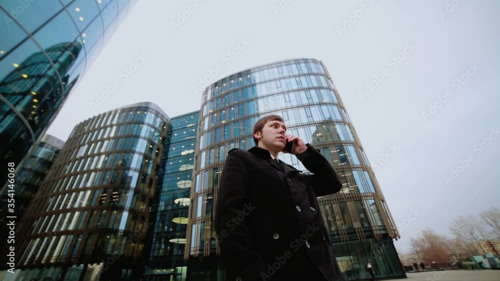 A male businessman in a black coat is talking on the phone in the street at a business center. A male businessman in a black coat is talking on the phone in the street at a business center.