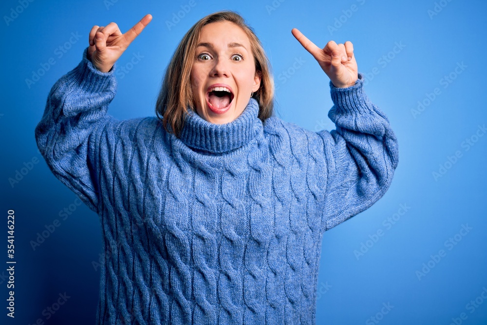 Young beautiful blonde woman wearing casual turtleneck sweater over blue background smiling amazed and surprised and pointing up with fingers and raised arms. Young beautiful blonde woman wearing casual turtleneck sweater over blue background smiling amazed and surprised and pointing up with fingers and raised arms.