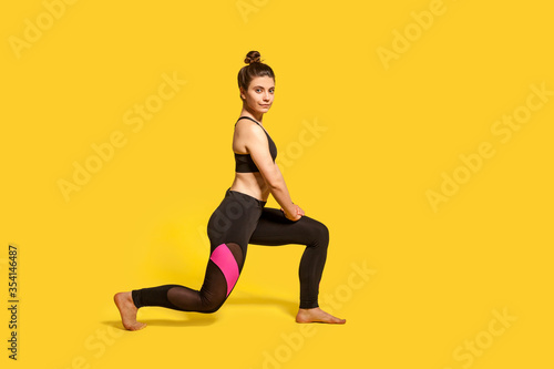 Athletic slim young woman with hair bun in tight sportswear doing sport lunge exercise, standing one knee, warming up training muscles, butts workout. full length studio shot, isolated on yellow © khosrork