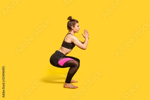 Athletic woman with hair bun in tight sportswear doing squat, lower body sport exercise, keeping balance, warming up and training muscles. full length studio shot, isolated on yellow background